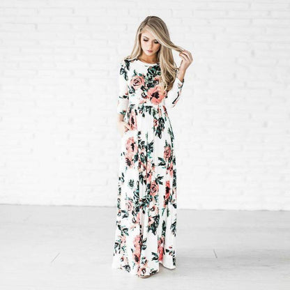 Frau in langem, geblümtem Kleid vor weißer Backsteinwand, modisches Outfit, florales Design, eleganter Look, Damenmode, stilvolle Kleidung.