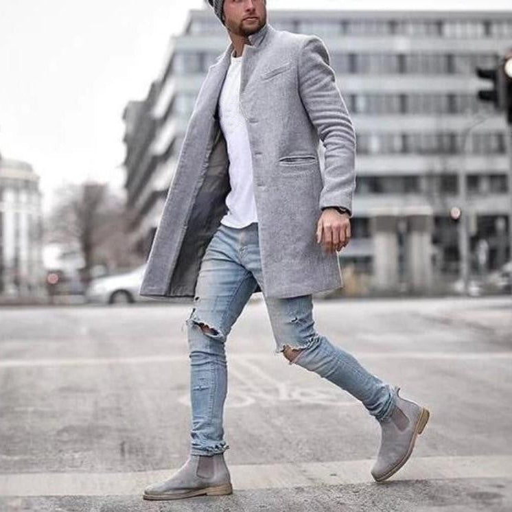 Mann in grauem Mantel, zerrissenen Jeans und Chelsea Boots überquert Straße in urbaner Umgebung. Modischer Streetstyle, Wintermode, Herrenbekleidung.