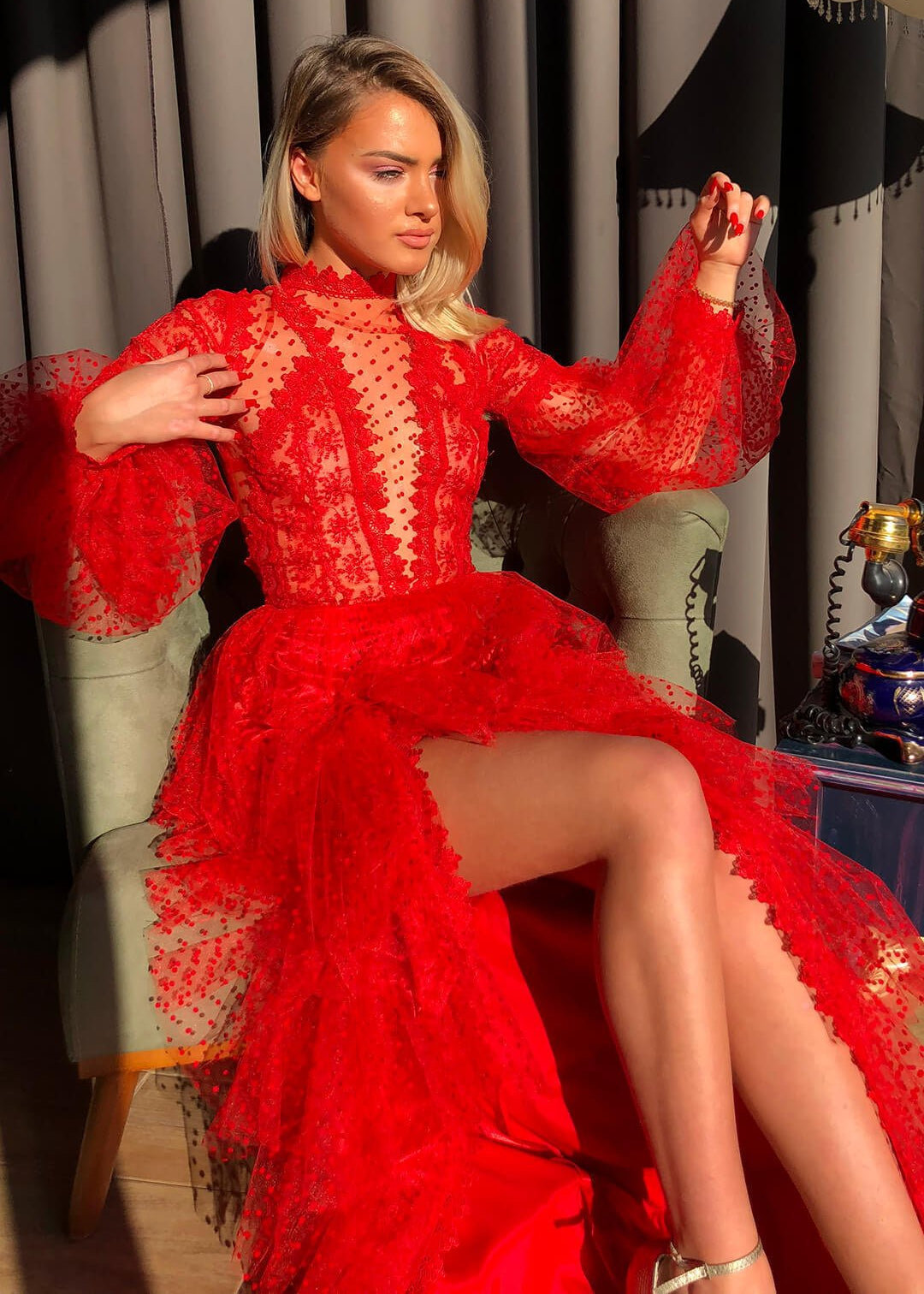 Frau in elegantem, rotem Spitzenkleid mit langen Ärmeln sitzt auf einem Sessel, Sonnenlicht betont das Design. Perfekt für Mode- und Abendkleider-Themen.