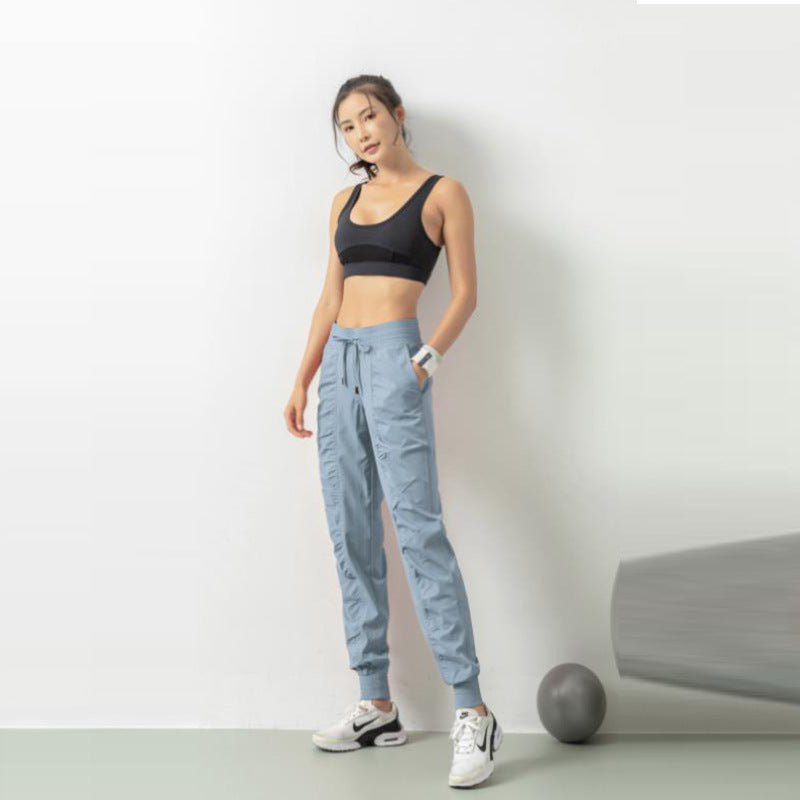 Frau in Sportbekleidung, schwarzes Top, blaue Jogginghose, weiße Sneaker, steht vor weißer Wand, Fitnessball am Boden, sportlich, modern.