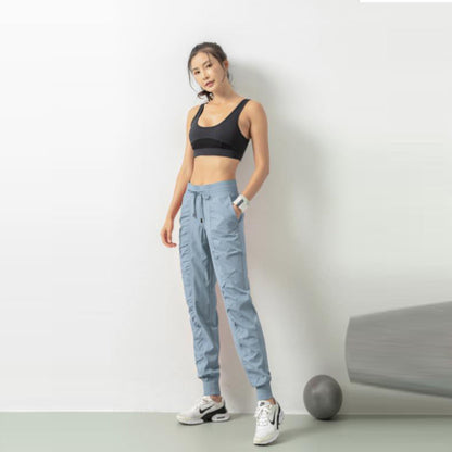 Frau in Sportbekleidung, schwarzes Top, blaue Jogginghose, weiße Sneaker, steht vor weißer Wand, Fitnessball am Boden, sportlich, modern.
