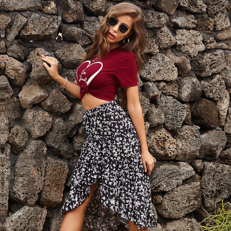 Frau in rotem T-Shirt mit Herzmotiv und schwarzem, geblümtem Rock vor Steinmauer. Modetrend, Sommeroutfit, lässiger Stil.