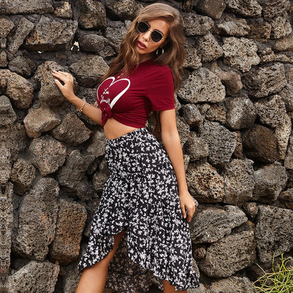 Frau in rotem T-Shirt mit Herzmotiv und schwarzem, geblümtem Rock vor Steinmauer. Modetrend, Sommeroutfit, lässiger Stil.