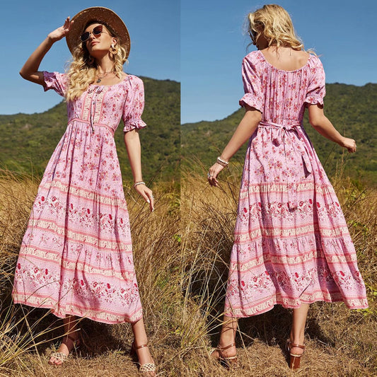Frau in rosa Sommerkleid mit Blumenmuster, Strohhut und Sonnenbrille steht in einer Wiese vor Berglandschaft. Boho-Chic, Mode, Outdoor.