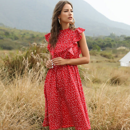 Frau in rotem, gepunktetem Kleid steht auf einer Wiese vor Berglandschaft. Sommermode, Freizeitkleidung, Naturhintergrund.