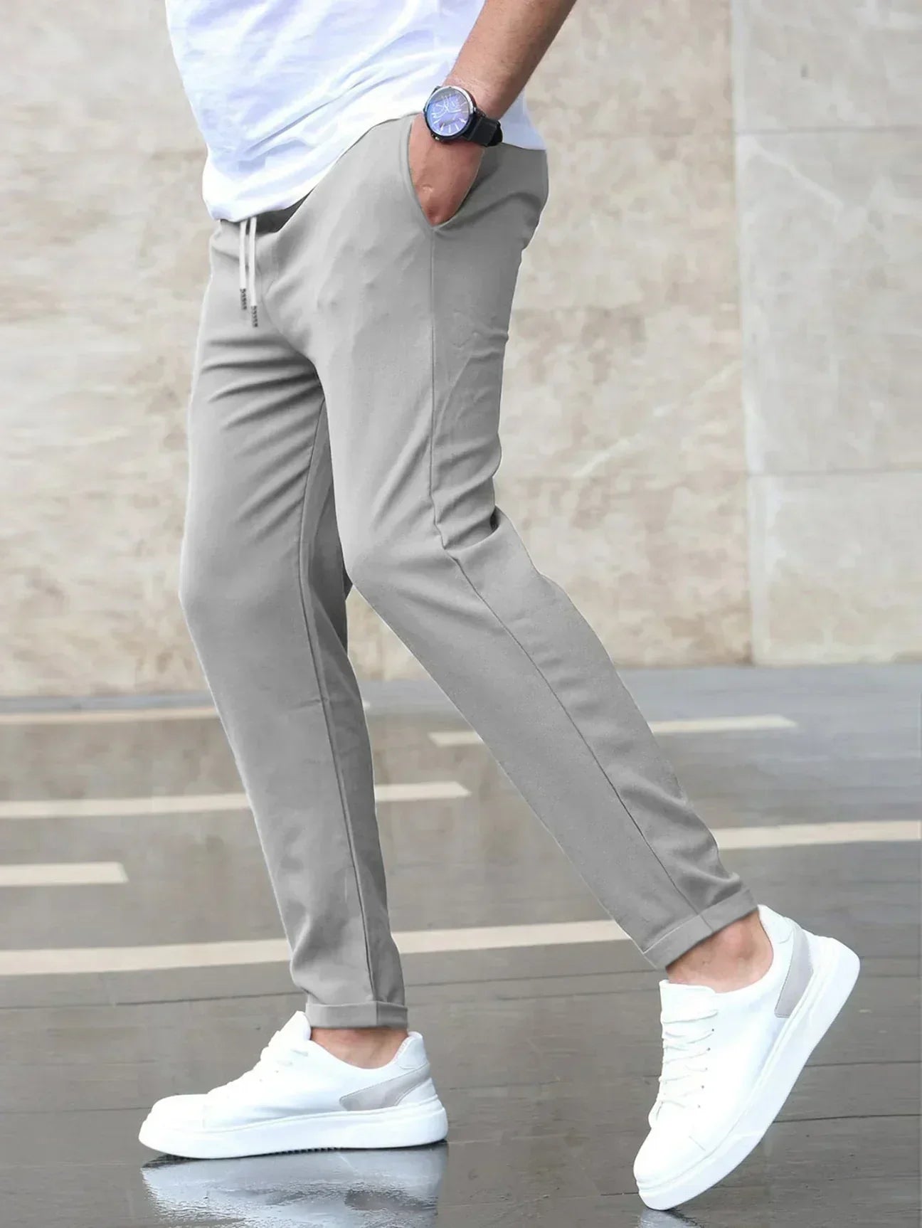 Mann in grauer Jogginghose und weißen Sneakers, trägt weißes T-Shirt, steht auf nassem Boden. Moderne Freizeitmode, Herrenbekleidung, stilvoll.