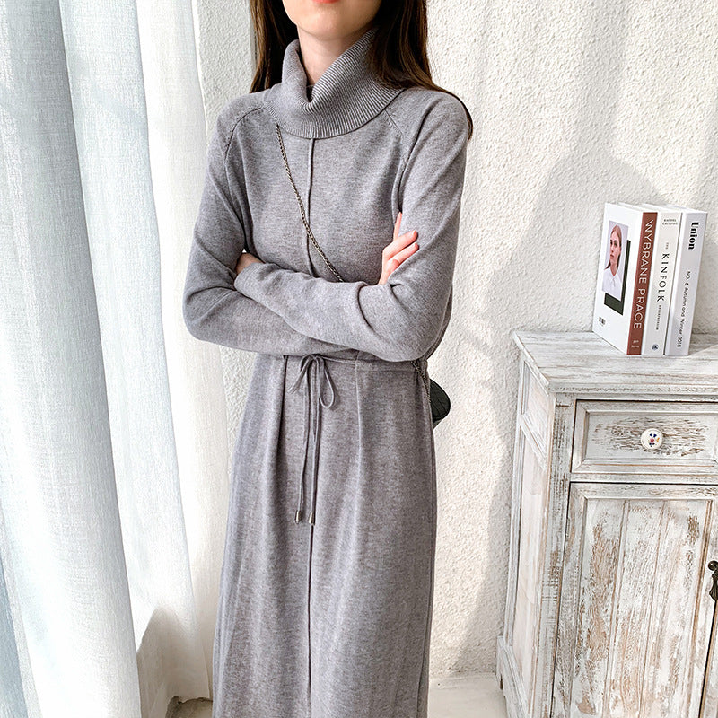 Frau in grauem Strickkleid mit Rollkragen, stehend neben einer weißen Kommode. Elegantes Winteroutfit, modisch und stilvoll.