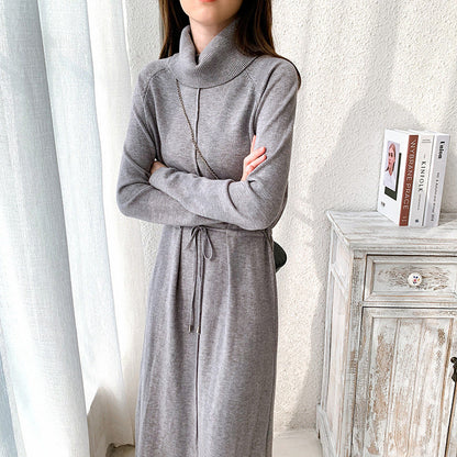 Frau in grauem Strickkleid mit Rollkragen, stehend neben einer weißen Kommode. Elegantes Winteroutfit, modisch und stilvoll.