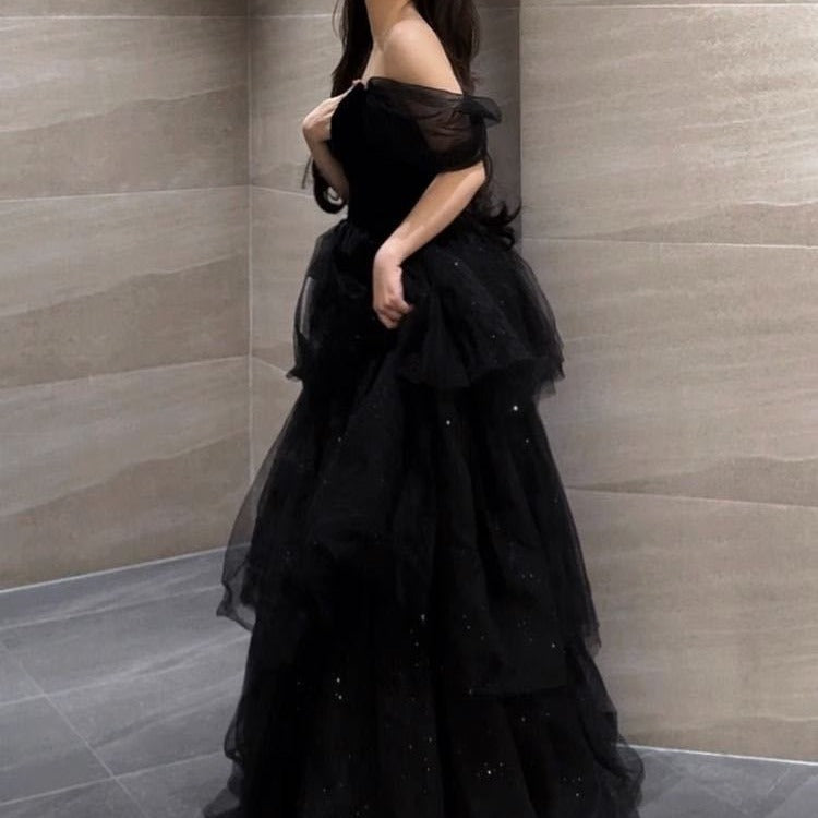Frau in elegantem, schwarzem Abendkleid mit schulterfreiem Design und Tüllrock, steht vor einer modernen, grauen Steinwand.