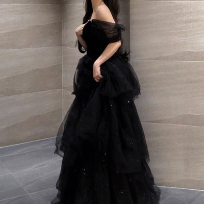 Frau in elegantem, schwarzem Abendkleid mit schulterfreiem Design und Tüllrock, steht vor einer modernen, grauen Steinwand.
