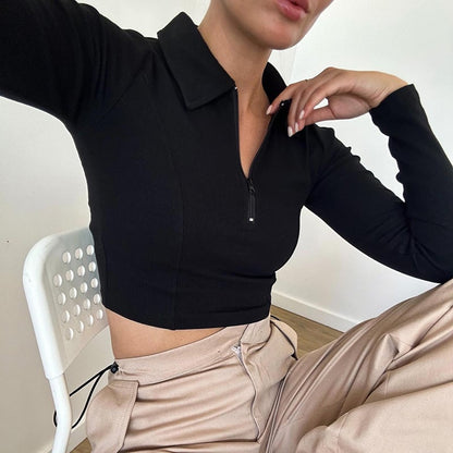Frau in schwarzem Langarm-Crop-Top und beigen Hosen sitzt auf weißem Stuhl. Modetrend, Freizeitkleidung, minimalistischer Stil.
