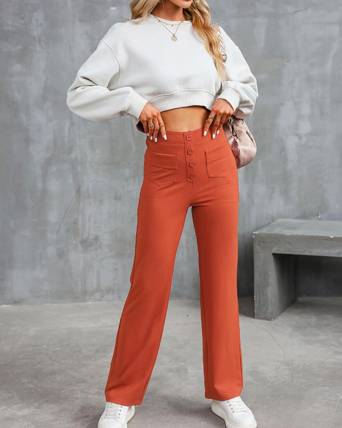 Frau in modischer, orangefarbener High-Waist-Hose und weißem Crop-Top vor grauer Wand, trägt weiße Sneaker und goldene Halskette.