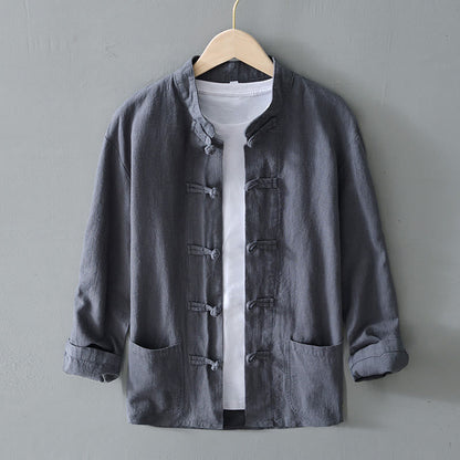 Graue Herrenjacke im chinesischen Stil mit Stehkragen und Knöpfen, über weißem T-Shirt, auf Holzkleiderbügel vor grauer Wand.