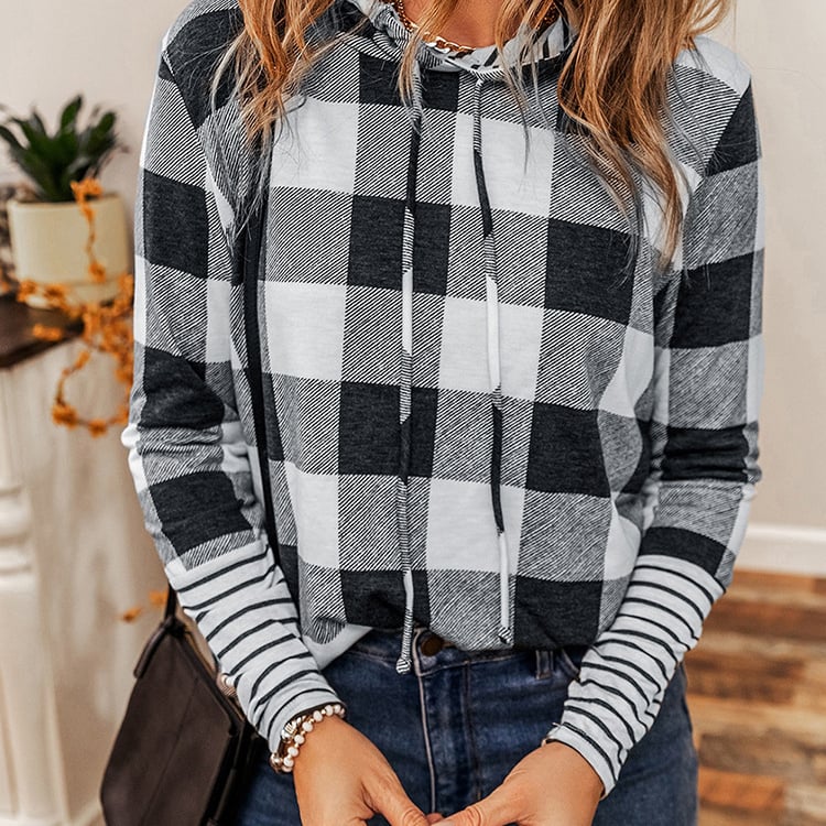 Frau in kariertem Kapuzenpullover und Jeans, modisch gekleidet, steht in modernem Raum. Trendige Herbstmode, lässiger Stil, gemütliches Outfit.