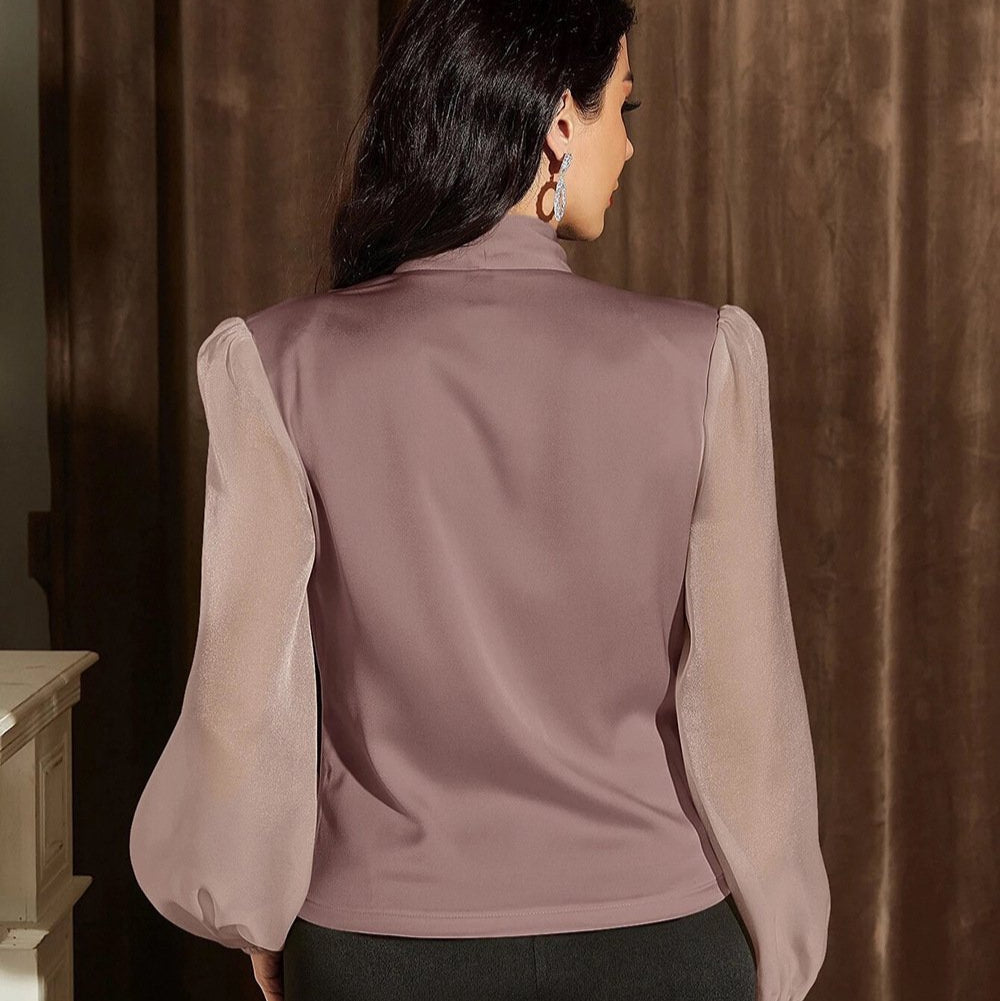 Frau in lila Bluse mit transparenten Ärmeln, steht vor braunem Vorhang. Modetrend, elegante Damenmode, stilvolle Bluse, Rückansicht.