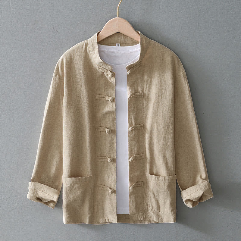 Beige Herrenjacke aus Leinen mit Stehkragen, Knopfleiste und aufgesetzten Taschen, über weißem T-Shirt auf Holzkleiderbügel vor grauer Wand.