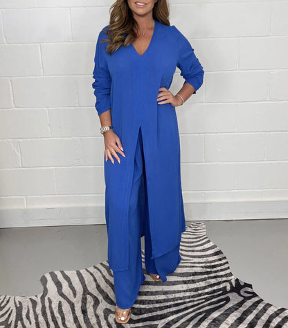 Frau in elegantem, blauem, langem Kleid mit V-Ausschnitt und Hose, steht auf einem Zebrateppich vor weißer Ziegelwand. Mode, Damenbekleidung, Stil.