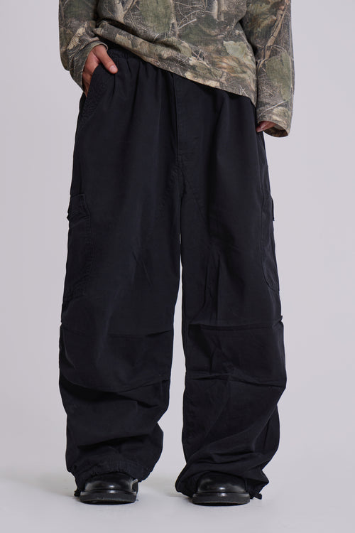 Person in schwarzer Baggy-Hose und Camouflage-Oberteil, trägt schwarze Schuhe. Streetwear, lässiger Stil, modische weite Hosen.