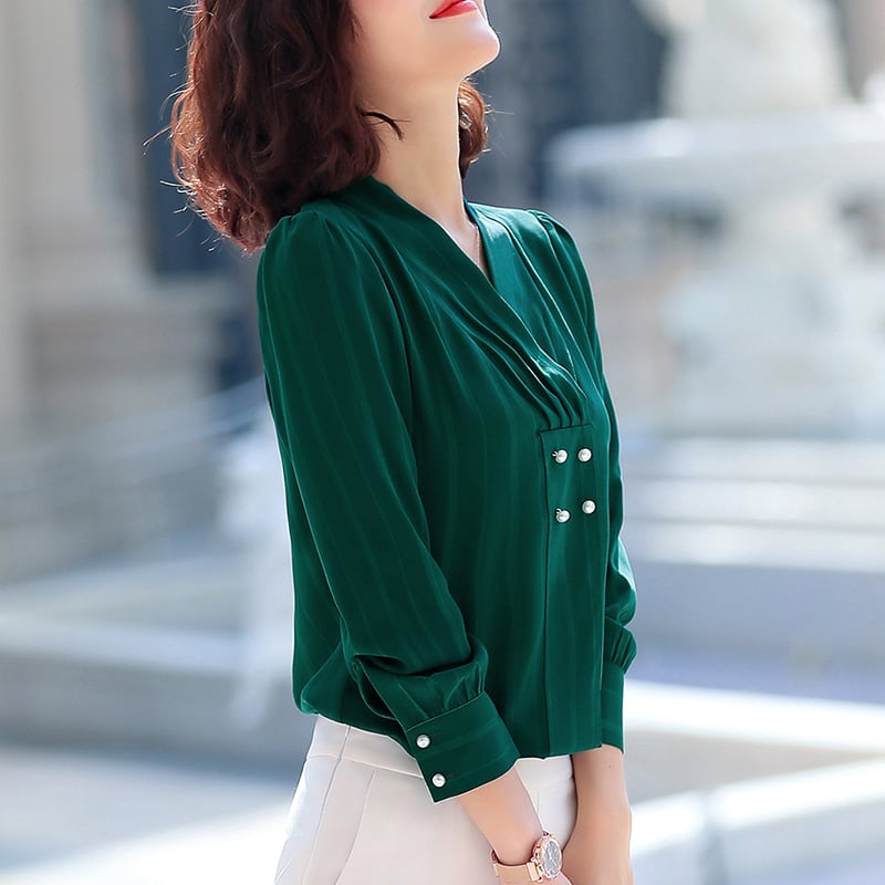 Frau in eleganter, grüner Bluse mit Perlenknöpfen, steht draußen. Modetrend, Damenmode, stilvolle Kleidung, urbaner Hintergrund.
