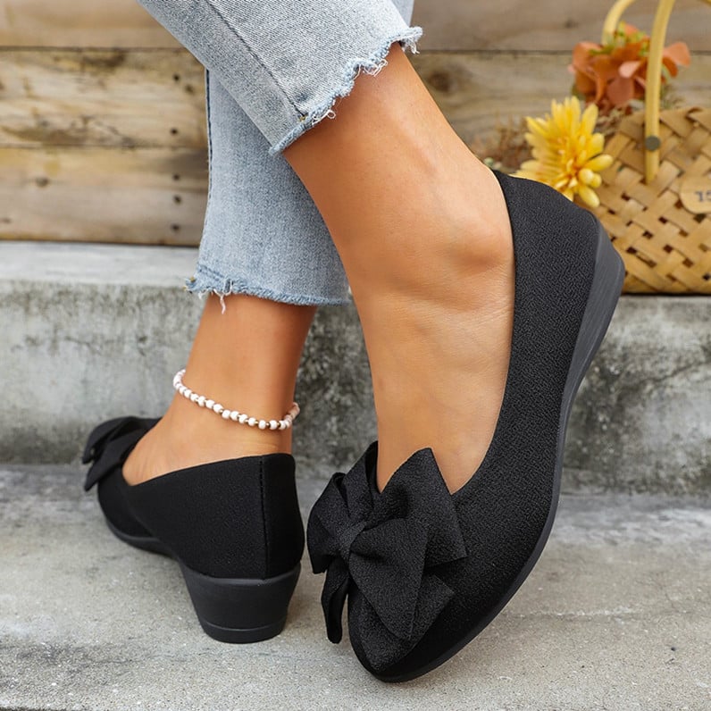 Schwarze Damen-Ballerinas mit Schleife, kombiniert mit hellblauen Jeans und Perlenfußkettchen, vor einem rustikalen Hintergrund mit Korb und Blumen.