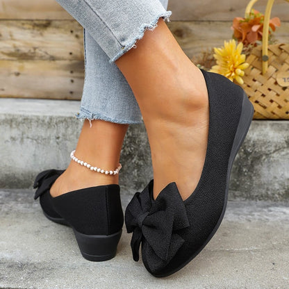 Schwarze Damen-Ballerinas mit Schleife, kombiniert mit hellblauen Jeans und Perlenfußkettchen, vor einem rustikalen Hintergrund mit Korb und Blumen.