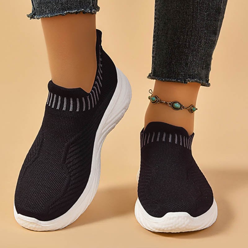 Schwarze Damen-Sneaker mit weißer Sohle, gestricktes Obermaterial, getragen mit Jeans und türkisfarbenem Fußkettchen. Bequeme Freizeitschuhe.