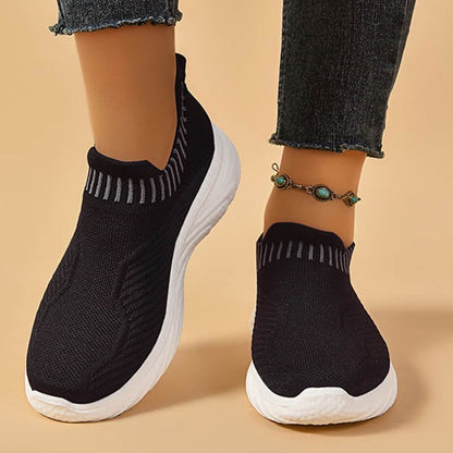 Schwarze Damen-Sneaker mit weißer Sohle, gestricktes Obermaterial, getragen mit Jeans und türkisfarbenem Fußkettchen. Bequeme Freizeitschuhe.