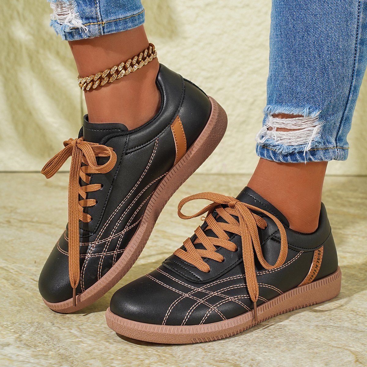 Schwarze Sneaker mit braunen Schnürsenkeln, getragen von Person in zerrissenen Jeans und goldener Fußkette. Modische Freizeitschuhe, Damenmode.