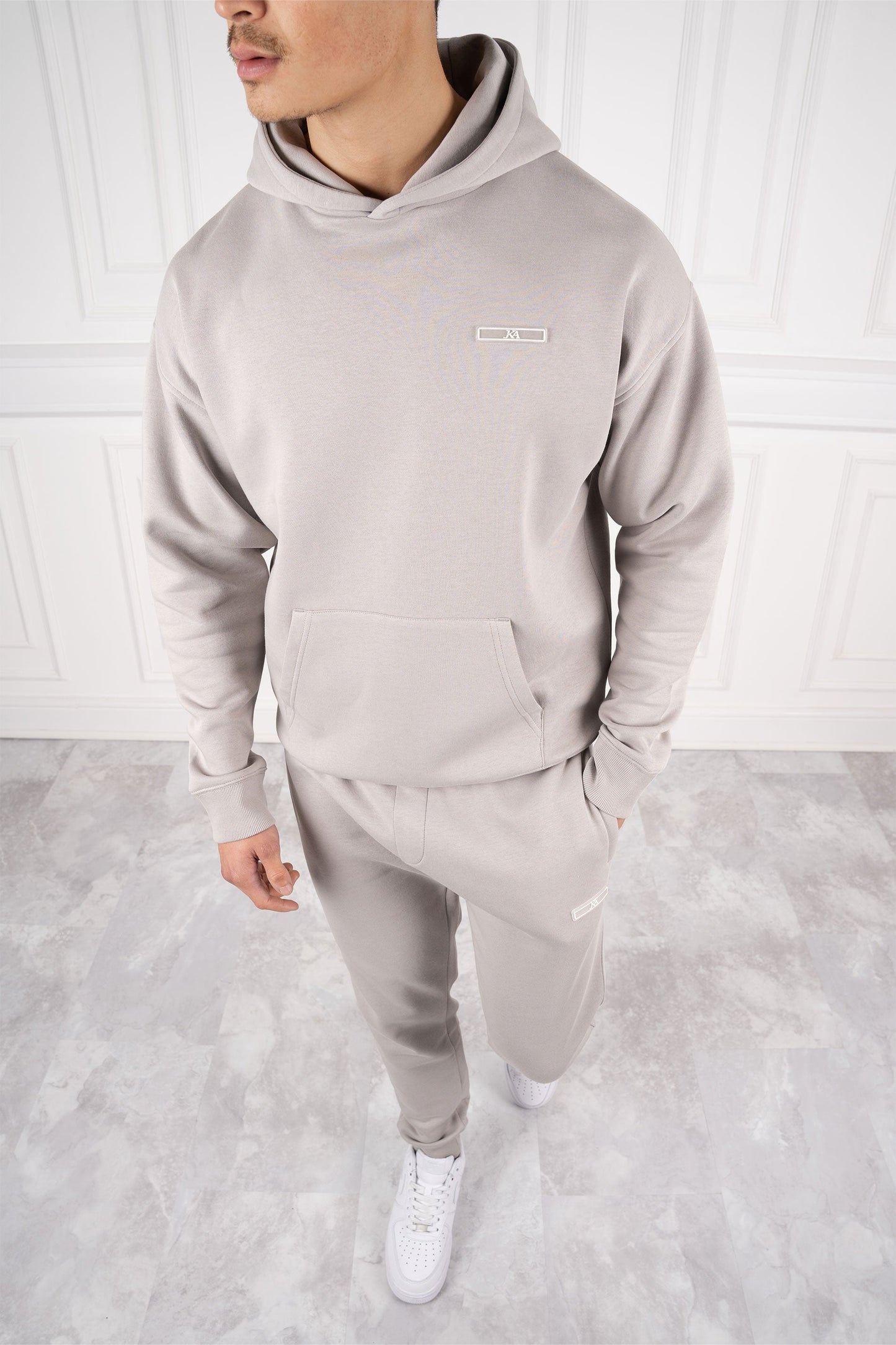 Mann in grauem Hoodie und Jogginghose, steht auf hellem Marmorboden, weiße Sneakers, moderne Freizeitmode, stilvoller Look.