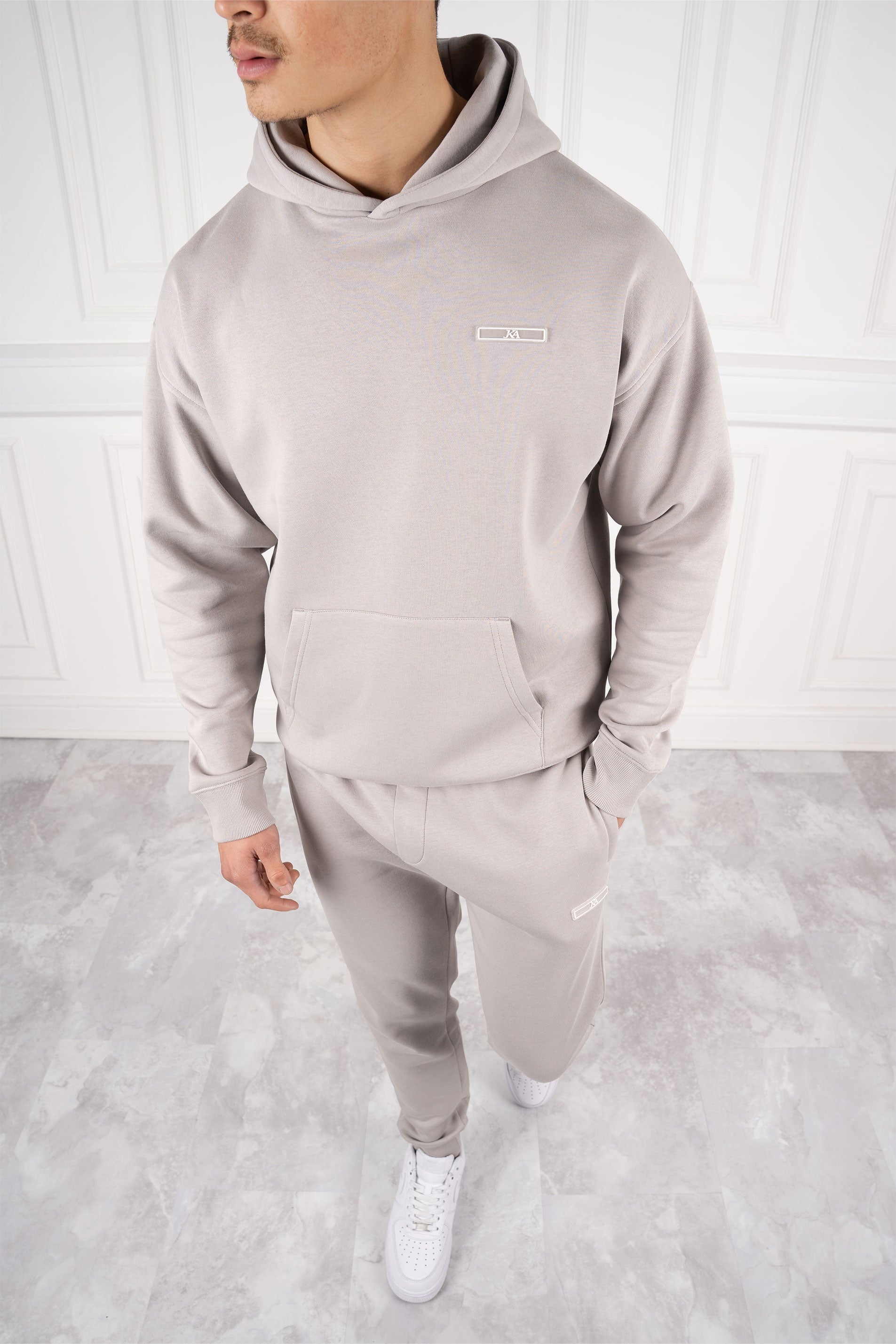 Mann in grauem Hoodie und Jogginghose, steht auf hellem Marmorboden, weiße Sneakers, moderne Freizeitmode, stilvoller Look.
