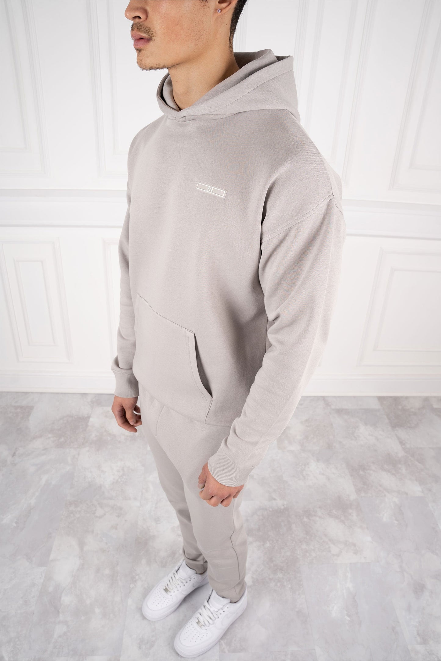 Mann in grauem Hoodie und Jogginghose, steht auf hellem Marmorboden, trägt weiße Sneaker. Moderne Herrenmode, Freizeitlook, Streetwear.