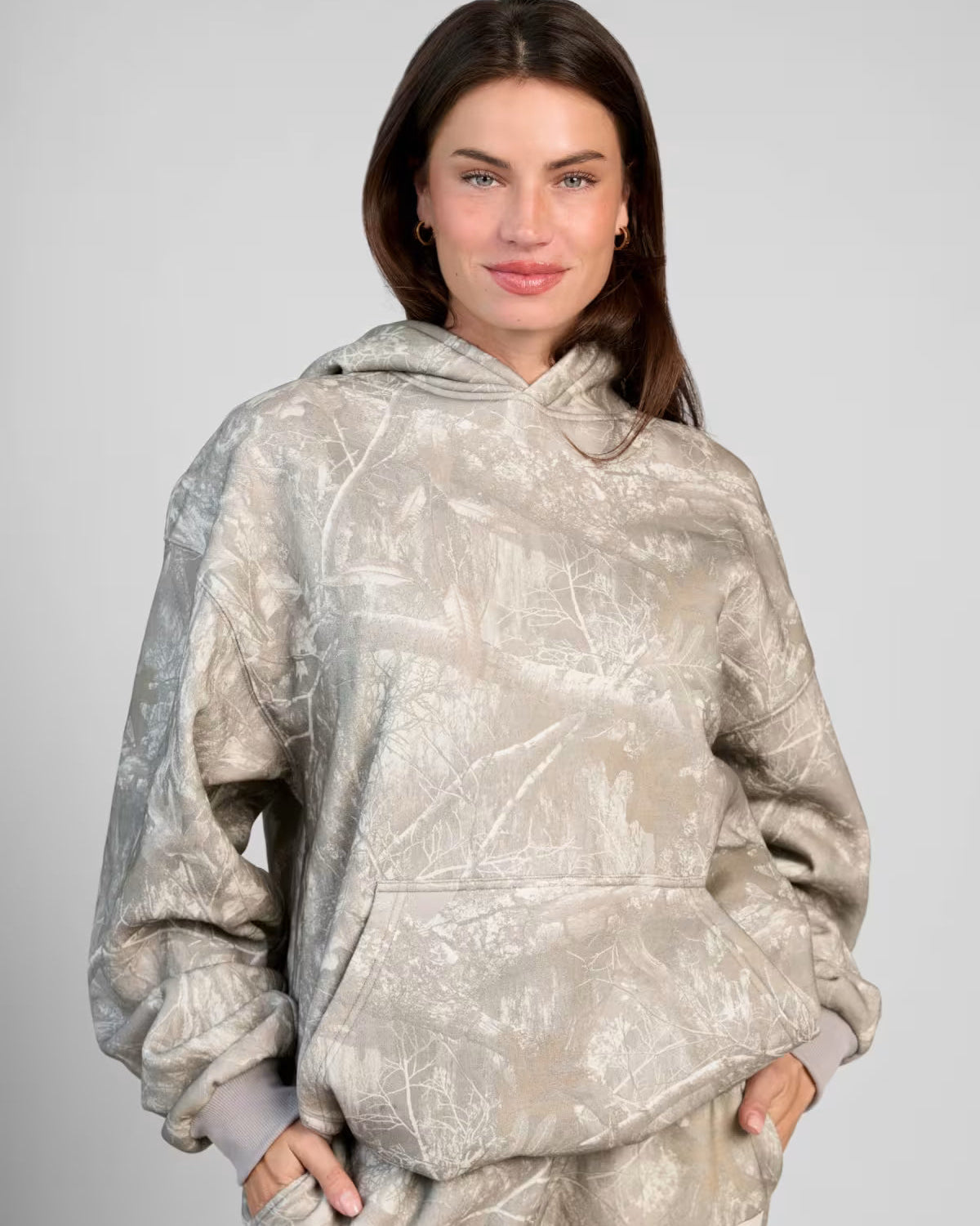 Frau in grauem Camouflage-Hoodie, lächelnd, mit Händen in den Taschen. Modetrend, lässiger Stil, Damenbekleidung, Herbstmode.