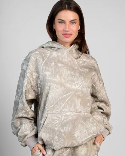 Frau in grauem Camouflage-Hoodie, lächelnd, mit Händen in den Taschen. Modetrend, lässiger Stil, Damenbekleidung, Herbstmode.