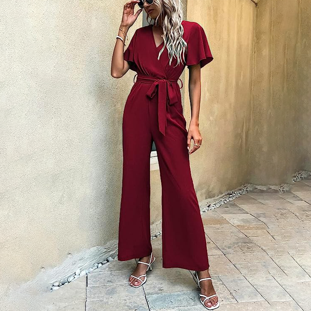 Frau in rotem Jumpsuit mit Gürtel, trägt Sonnenbrille und Sandalen, steht auf gepflastertem Boden. Elegante Damenmode, Sommeroutfit, stilvoll.