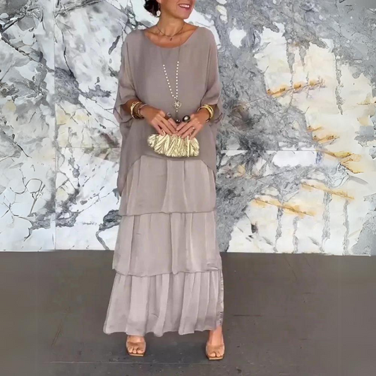 Elegantes Outfit mit langem, fließendem Kleid in Beige, goldener Clutch und Schmuck, vor marmorierter Wand. Perfekt für stilvolle Anlässe.