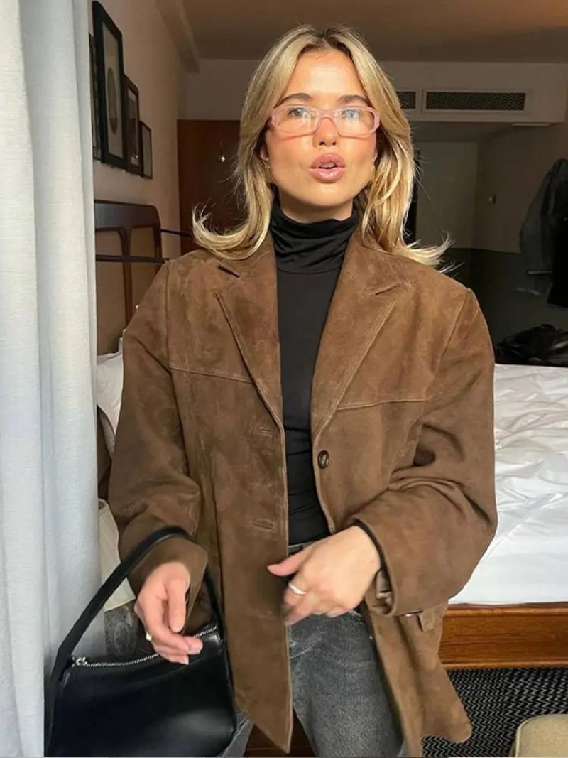 Frau mit brauner Wildlederjacke und Brille steht in modernem Schlafzimmer, hält schwarze Handtasche. Modetrend, Herbstmode, stilvolle Kleidung.