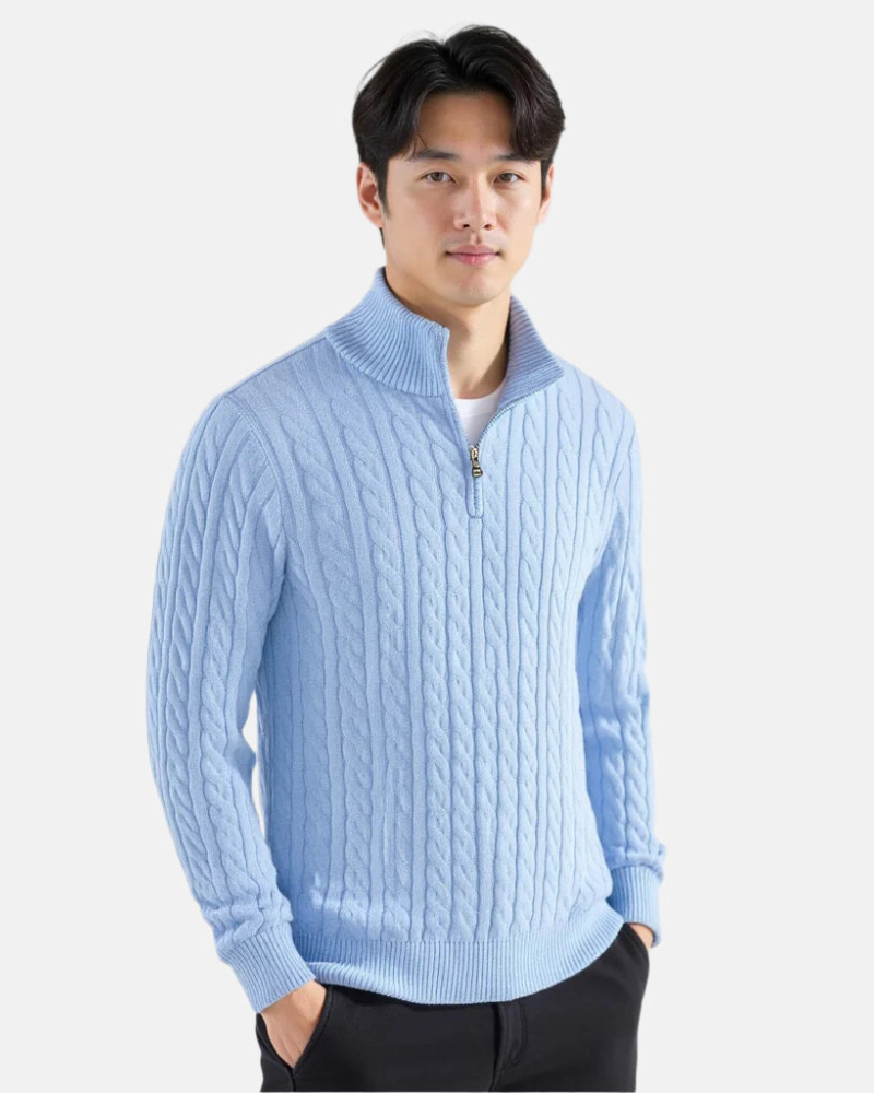 Ein Mann trägt einen hellblauen Zopfmuster-Pullover mit Reißverschlusskragen, kombiniert mit schwarzen Hosen. Elegante Herrenmode, Winteroutfit.