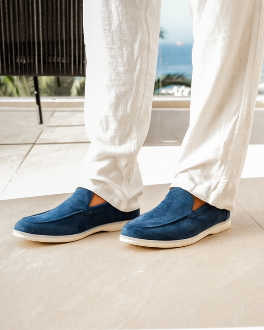 Weiße Leinenhose und blaue Wildleder-Slipper auf heller Terrasse mit Meerblick. Elegante Sommermode, Herren-Schuhe, stilvolle Freizeitkleidung.