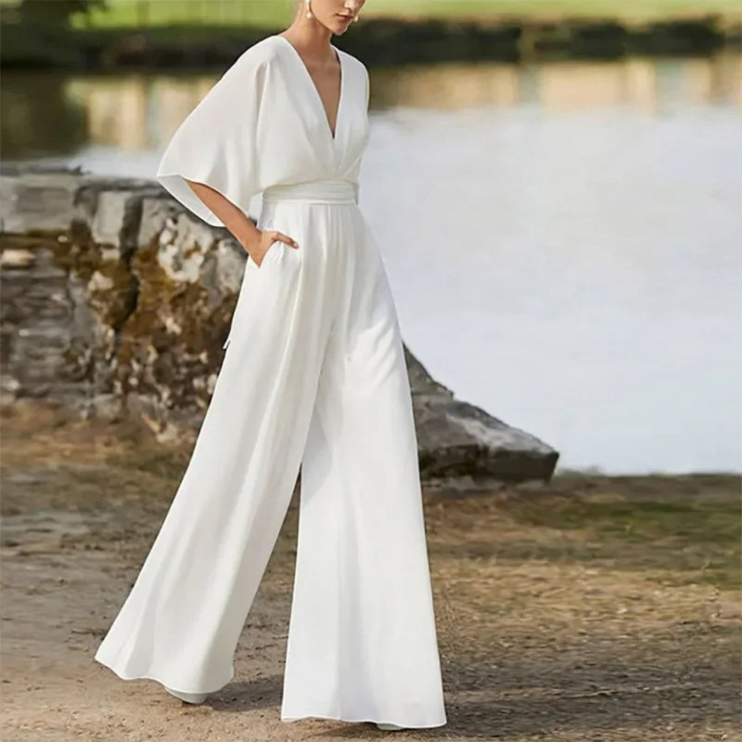 Frau in elegantem weißen Jumpsuit mit weitem Bein und tiefem V-Ausschnitt, steht draußen vor einem See. Ideal für Sommermode und elegante Anlässe.
