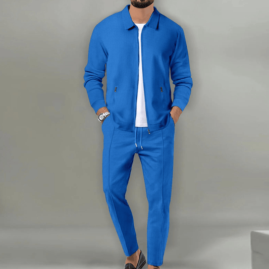 Mann in blauem Anzug mit weißem T-Shirt und Uhr, lässig posierend vor grauem Hintergrund. Elegante Herrenmode, stilvoll und modern.