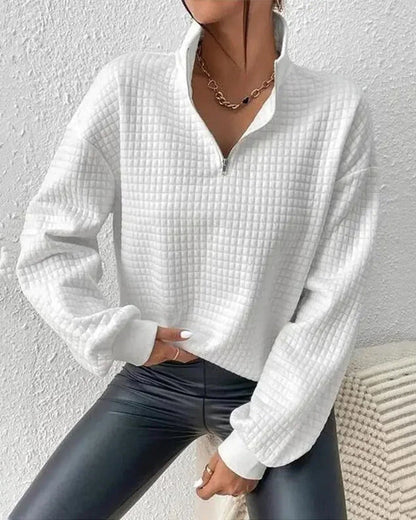Frau in weißem, gestepptem Pullover mit Reißverschluss und schwarzer Lederhose vor weißer Wand. Modetrend, Damenmode, Herbstoutfit.