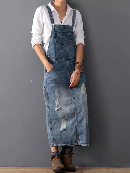 Frau in zerrissenem Jeans-Latzkleid und weißem Hemd, lässiger Look, braune Stiefel, grauer Hintergrund. Modetrend, Streetstyle, Denim.