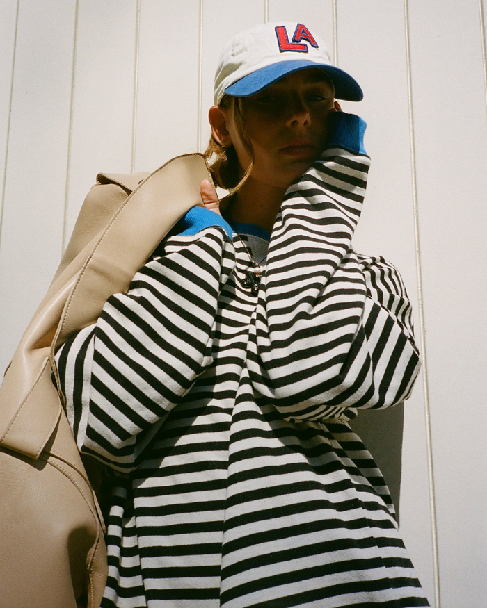 Frau in gestreiftem Pullover und LA-Cap vor weißer Wand, trägt beigefarbene Tasche. Streetwear, Mode, urbaner Stil, lässiger Look.