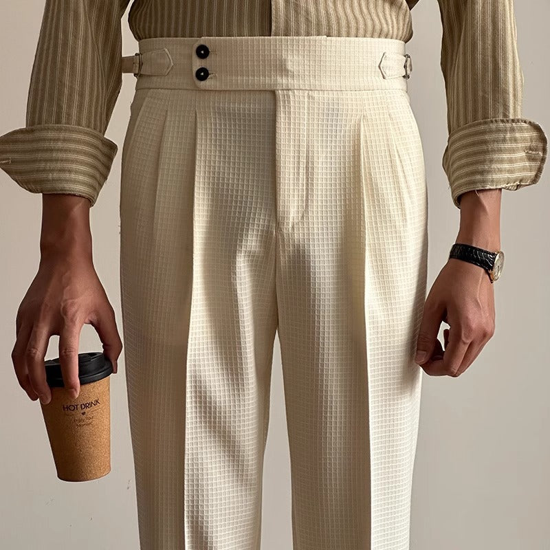 Mann in cremefarbener, karierter Anzughose mit Kaffeebecher. Elegante Herrenmode, Business-Look, gestreiftes Hemd, stilvolles Accessoire.