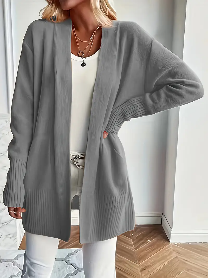 Frau in grauem Strick-Cardigan, weißem Top und Hose, steht in modernem Raum. Modetrend, lässiger Stil, Damenmode, Herbstoutfit.