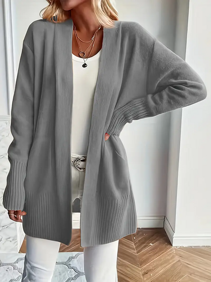 Frau in grauem Strick-Cardigan, weißem Top und Hose, steht in modernem Raum. Modetrend, lässiger Stil, Damenmode, Herbstoutfit.