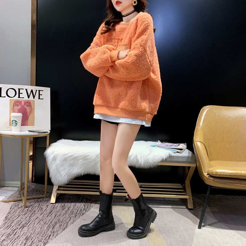 Frau in orangefarbenem Oversize-Pullover und schwarzen Stiefeln, steht in modernem Raum mit Loewe-Poster, weißem Fell und beigem Sessel.