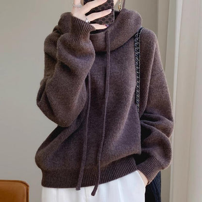 Frau in braunem Oversize-Pullover mit Kapuze und Kordeln, hält Smartphone. Modetrend, gemütlicher Strick, Herbst-Winter-Outfit.
