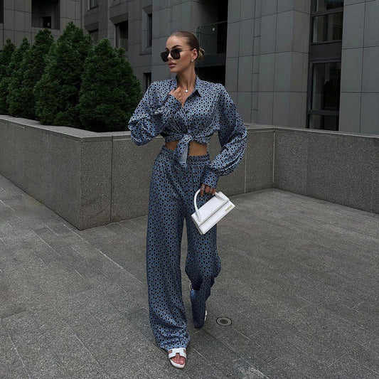 Frau in blauem gemustertem Outfit mit weißer Handtasche und Sonnenbrille, steht vor modernem Gebäude. Elegante Mode, urbane Kulisse.