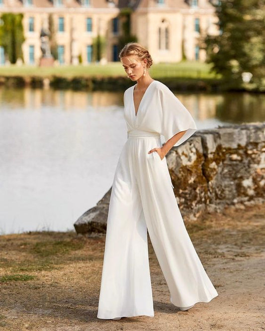 Frau in elegantem weißen Jumpsuit mit weitem Bein, steht vor einem historischen Gebäude und See, stilvolle Hochzeitsmode, Sommerhochzeit.